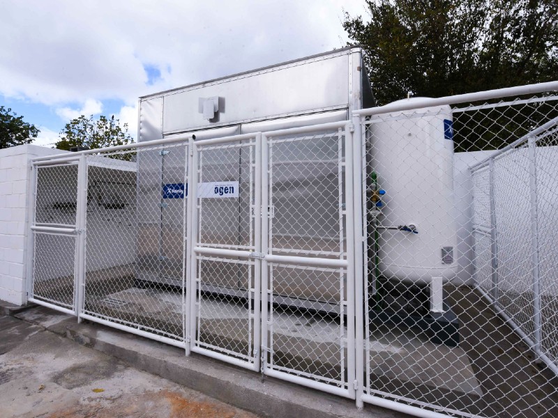 Foto da miniusina de oxigênio da UPA Jabaquara, composta por um compartimento metálico retangular e cilindro. A miniusina está cercada por um portão de ferro branco, que está fechado